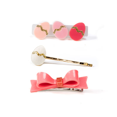 Trio of hair accessories. Two of them are hair clips and one is a bobby pin. The bobby pin is adorned with one pearlized easter egg. One of the hair clips is adorned with a pink bow and the other one has three easter eggs. 